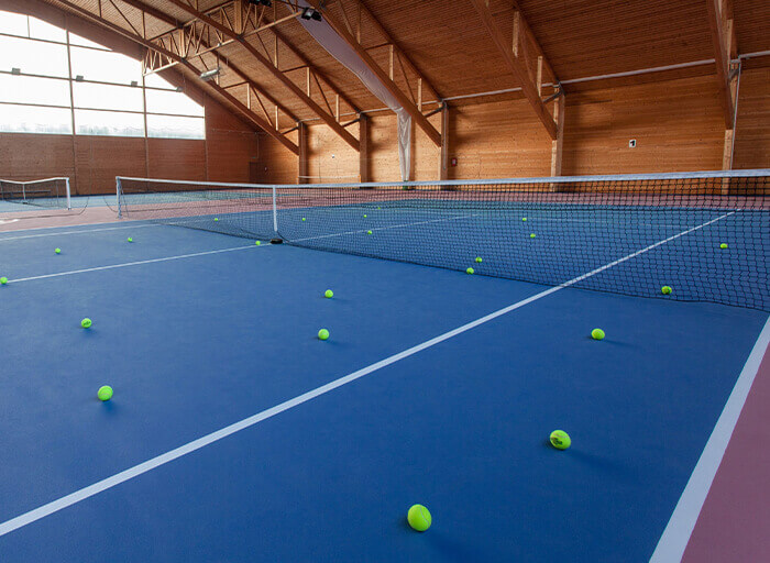 Modrý tenisový kurt s rozesetými míčky ve vnitřní dřevěné hale.